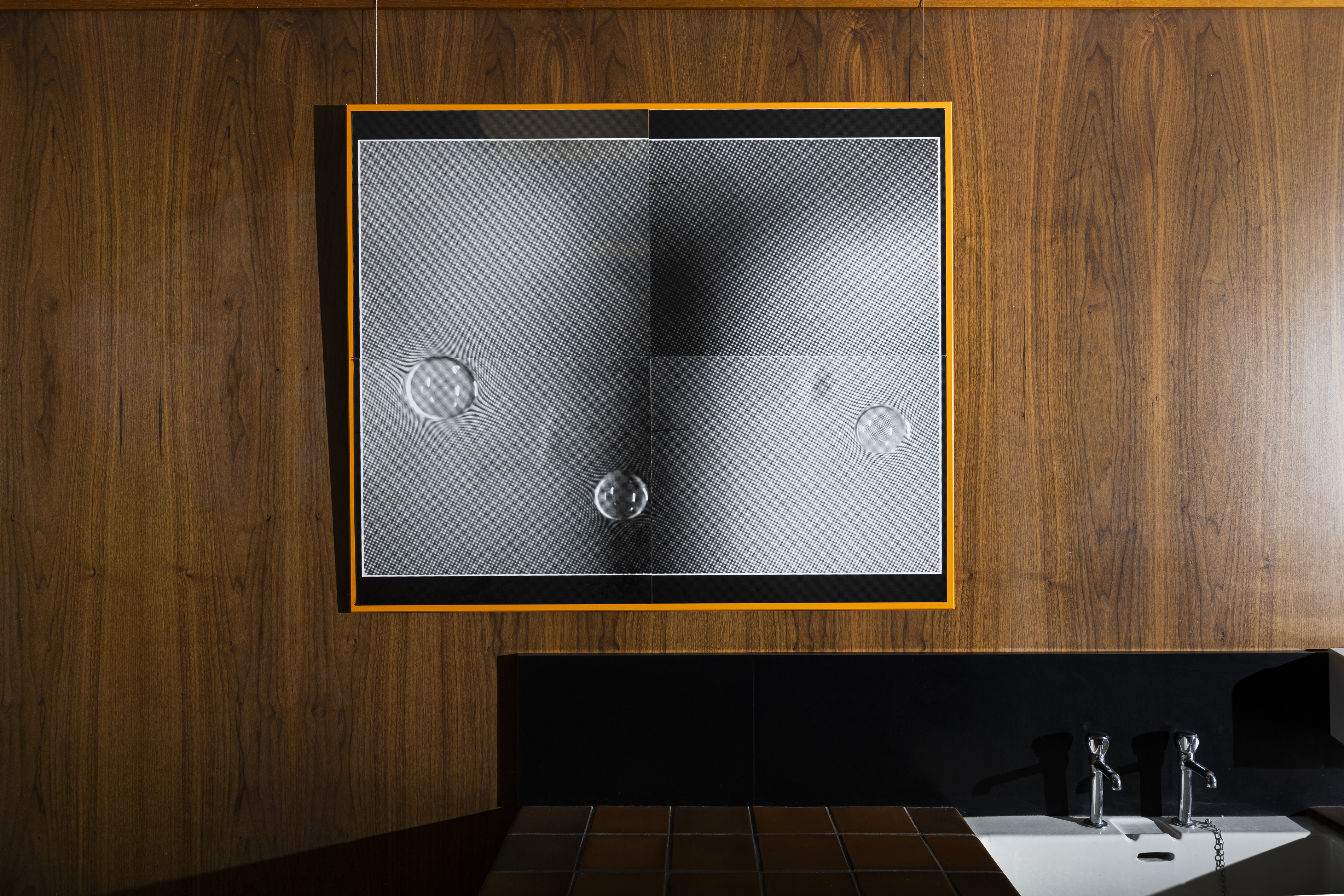 Orange-framed artwork on wood panelling showing a high-contrast halftone pattern with three glass-like bubbles, hung above a black-tiled sink with chrome taps.