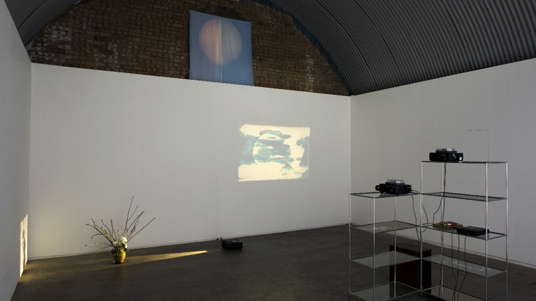 white wall in a brick space with a metal corrugated roof. there is a vase on the floor illuminated by a beam of yellow light. a projection on the wall of a red circle on a yellow and green background and two metal and glass shelves. 
