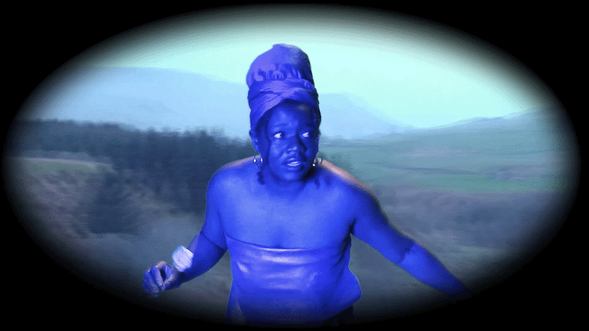 A woman, painted all in blue, wearing a turban and a strapless top looks to the side. The background is of a wooded landscape.