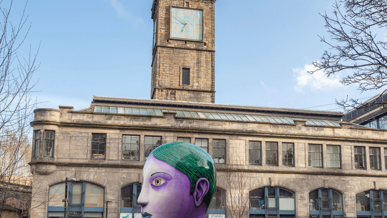 Profile right view of a big, tree-sized bust of a woman with purple skin, green eyes, thin eyebrows and green hair and lips. It is positioned in a park.