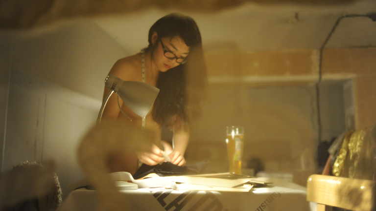 Nude person working at a dimly lit table with papers, under a desk lamp, with a drink nearby. They are wearing glasses, a measuring tape around their neck and focused on the task