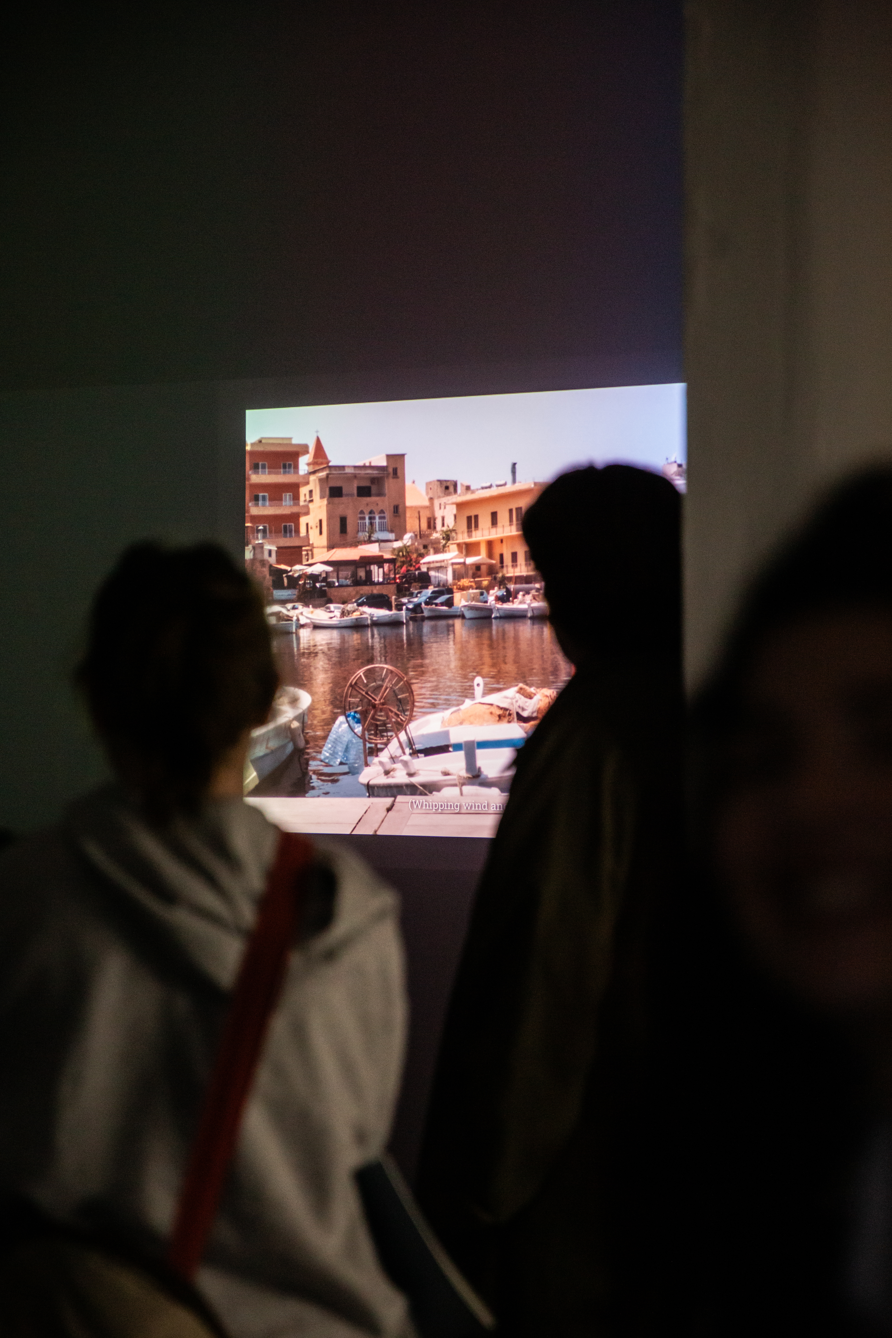 Visitors silhouetted in front of a film projection showing a sunlit harbour lined with waterfront buildings and moored boats, with subtitled dialogue visible on screen.