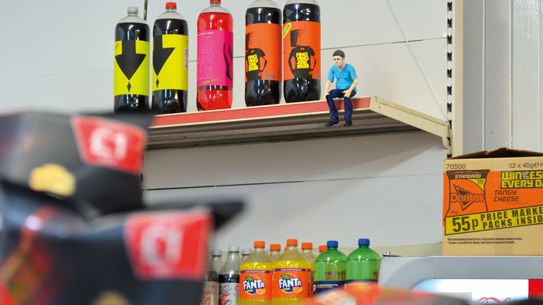 In a department store, there are drinks displayed. In the top shelf there are several colourful bottles with labels. Beside it, a small plastic figurine of a white man with blue t-shirt and jeans. 