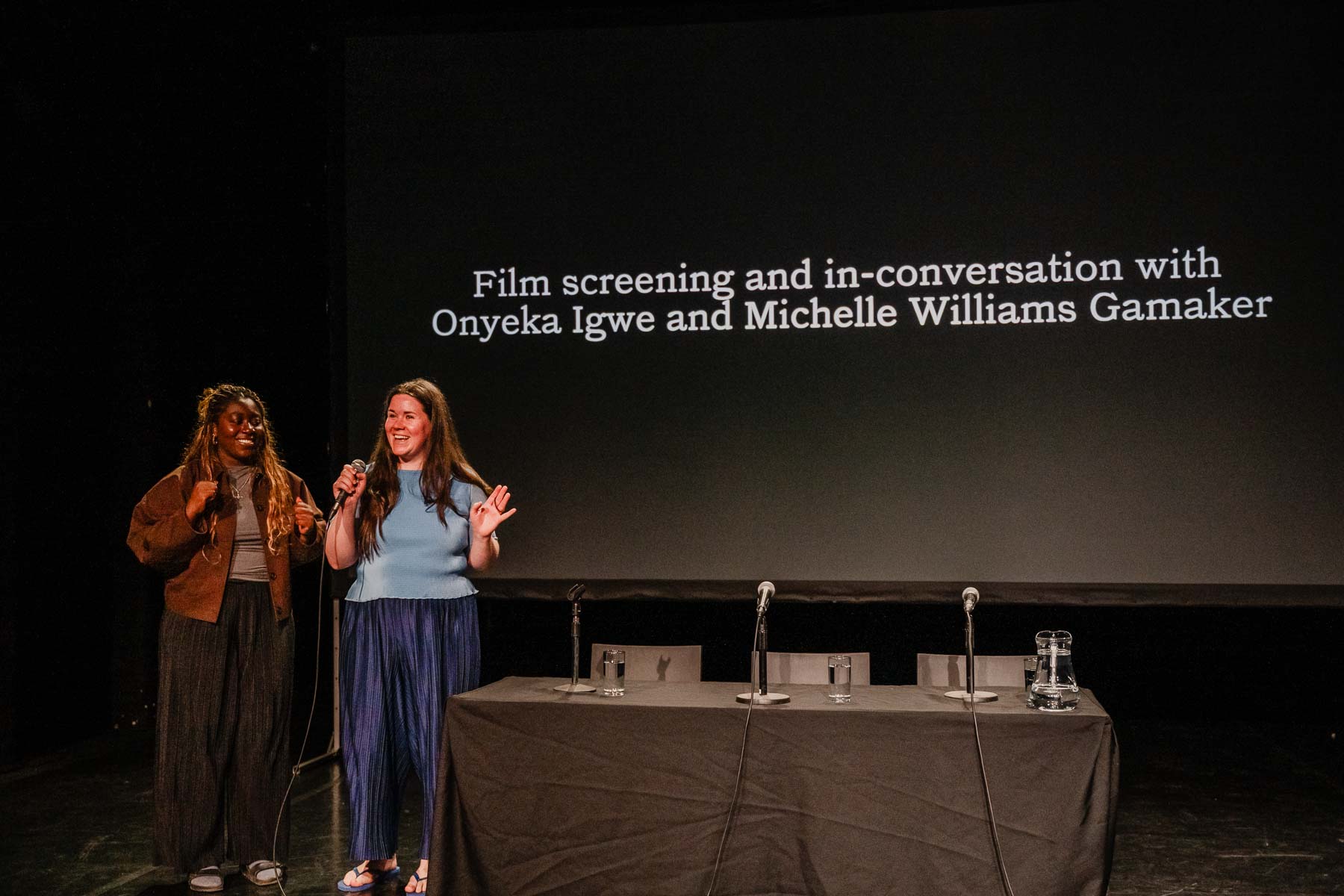 Two women stand smiling on a dark stage holding a microphone, in front of a projected screen reading "Film screening and in-conversation with Onyeka Igwe and Michelle Williams Gamaker." An empty panel table with microphones and water glasses sits in front of them.