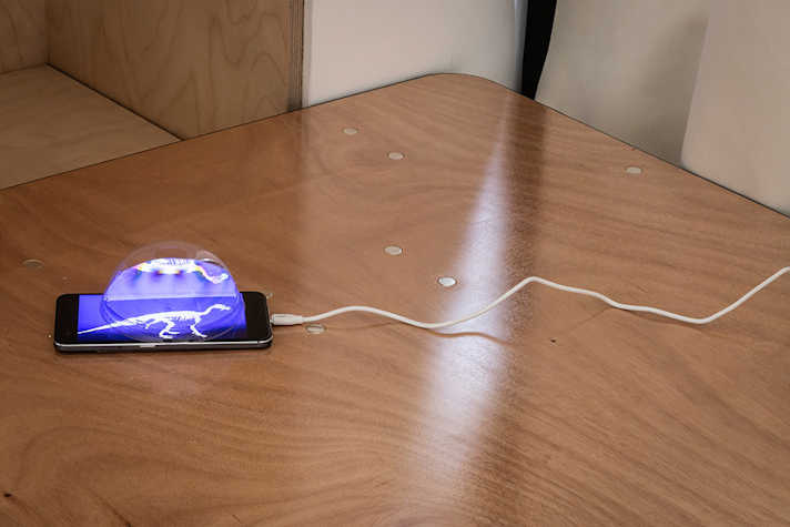 A phone displaying an image of a dinosaur is displayed on a table with a cable connected to it. There is a spherical see-through shape on top of the phone screen.