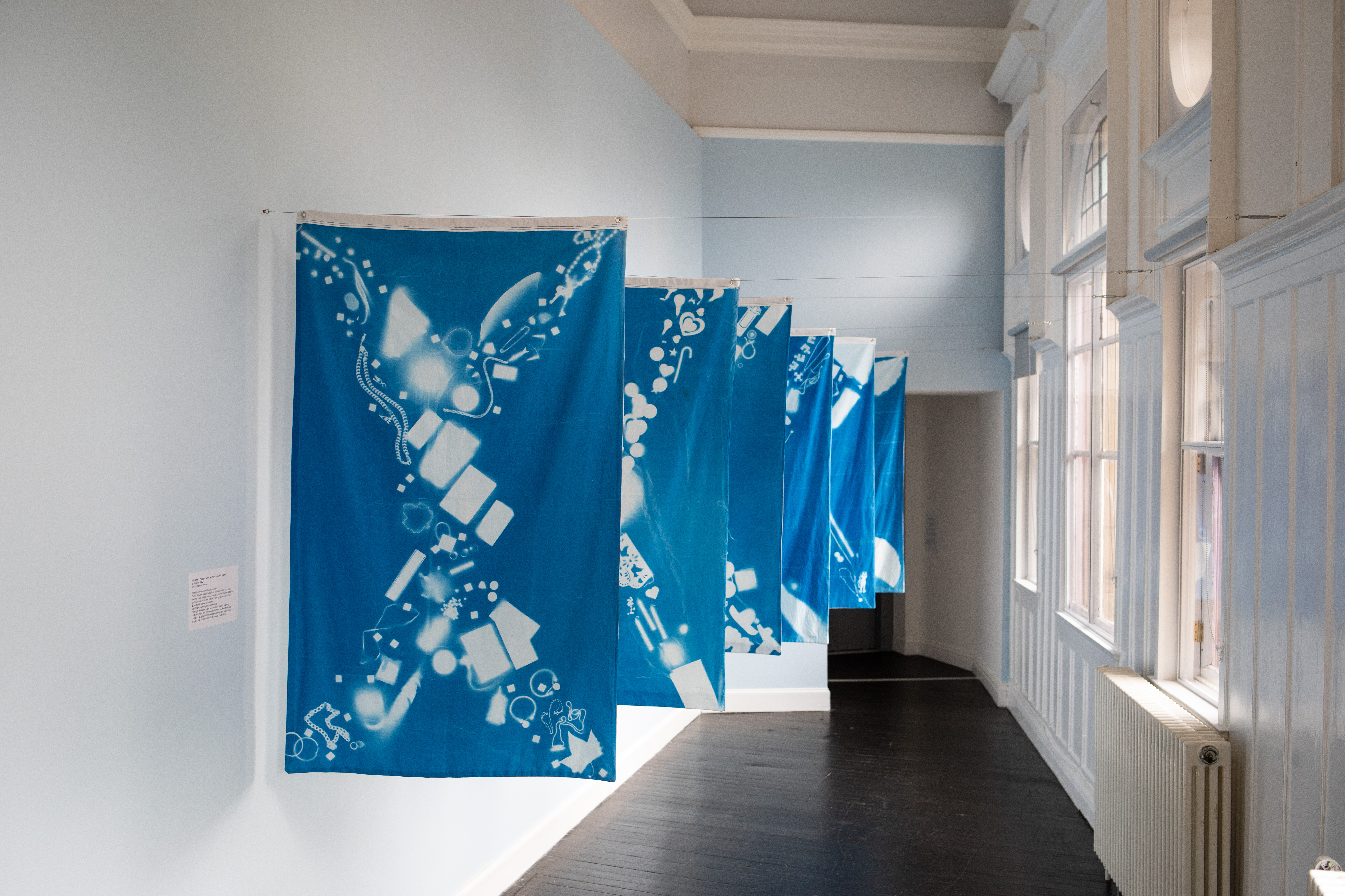 A row of hanging cyanotype banners in deep blue, each printed with white silhouettes of everyday objects like jewellery, buttons, and small trinkets, lit by tall windows along a gallery corridor.
