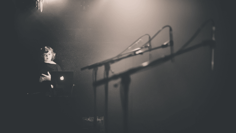 In a dark room, with some spotlights, there are three microphone stands standing next to each other. At the back of the room, there is a woman looking at their laptop.