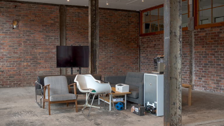 A casual seating area within a stripped-back brick interior, with a sofa, armchairs, coffee table, television and a small appliance, steel columns supporting the space.