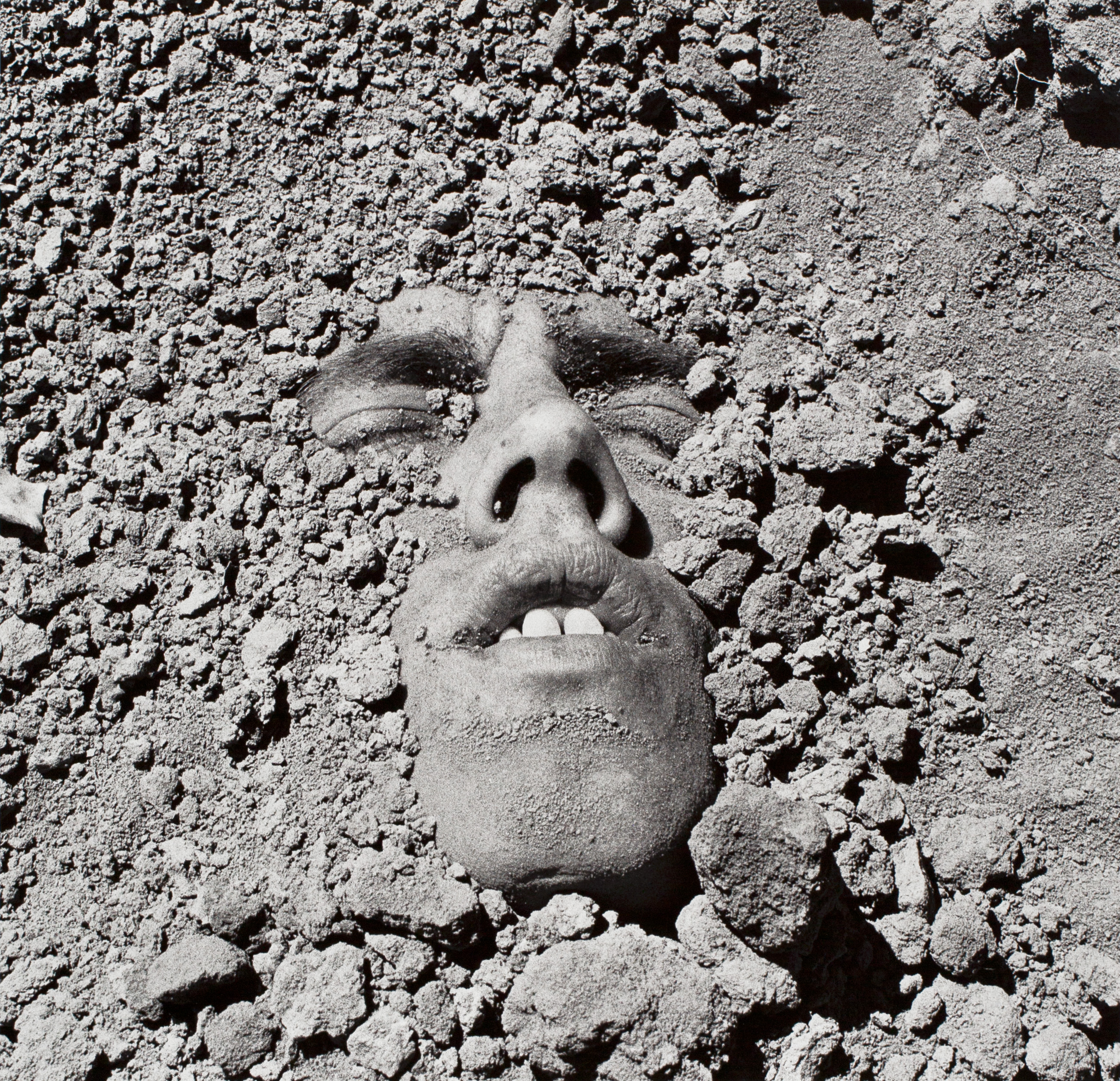 A black and white photograph of a person's face emerging  from the ground as if they have been buried. They are partially  covered in dirt, their eyes are closed and they are frowning slightly.