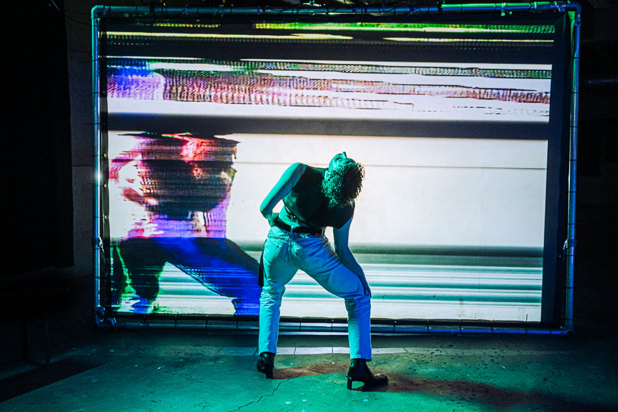 A performer bends forward dramatically in silhouette against a large screen of glitching static, under green stage lighting.