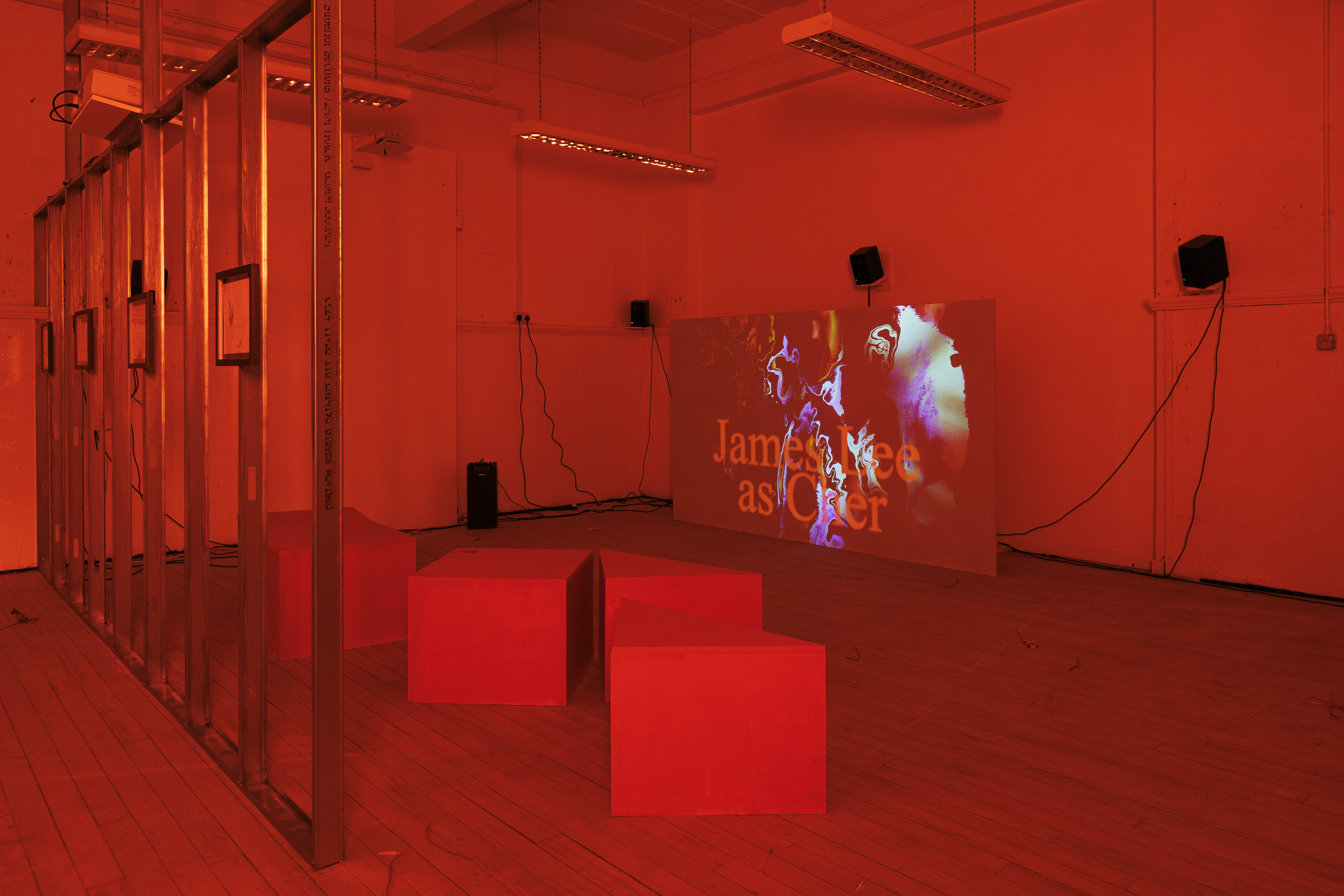 A room is shown saturated in red, as if seen through a red filter. On the left of the image, a metal frame wall is visible bearing four framed drawings. In the centre of the room are four square and triangular seats, and on the right is a projection screen. On the screen is a still image, the background of which shows abstract coloured shapes and in the foreground the words in large text: James Lee as Cher.