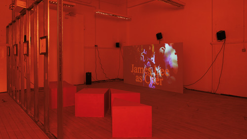 A room is shown saturated in red, as if seen through a red filter. On the left of the image, a metal frame wall is visible bearing four framed drawings. In the centre of the room are four square and triangular seats, and on the right is a projection screen. On the screen is a still image, the background of which shows abstract coloured shapes and in the foreground the words in large text: James Lee as Cher.