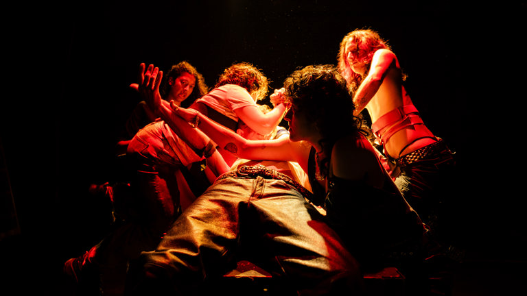 Five dyke-identifying peformers restrain and tought another performer lying on a table; only the performer's legs and crotch in jeans are visible. The scene is lit by red wash lighting in a dark space.