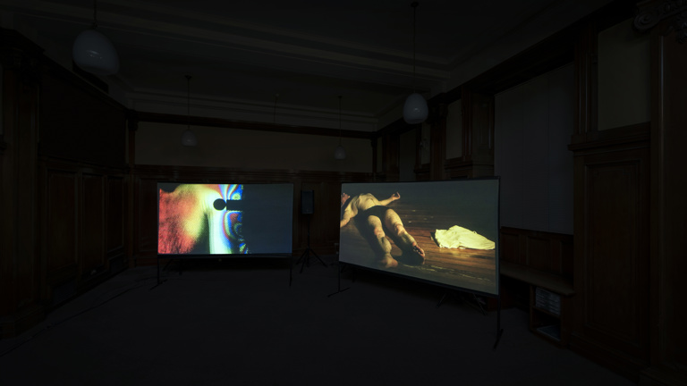 In a dark room two screens are forming a corner. The left screen has a holographic effect video still. The right screen has a man lying on the floor undressed. 