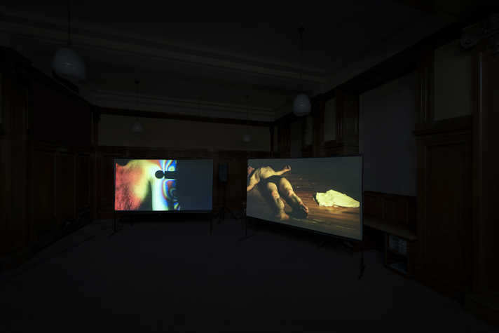 In a dark room two screens are forming a corner. The left screen has a holographic effect video still. The right screen has a man lying on the floor undressed.