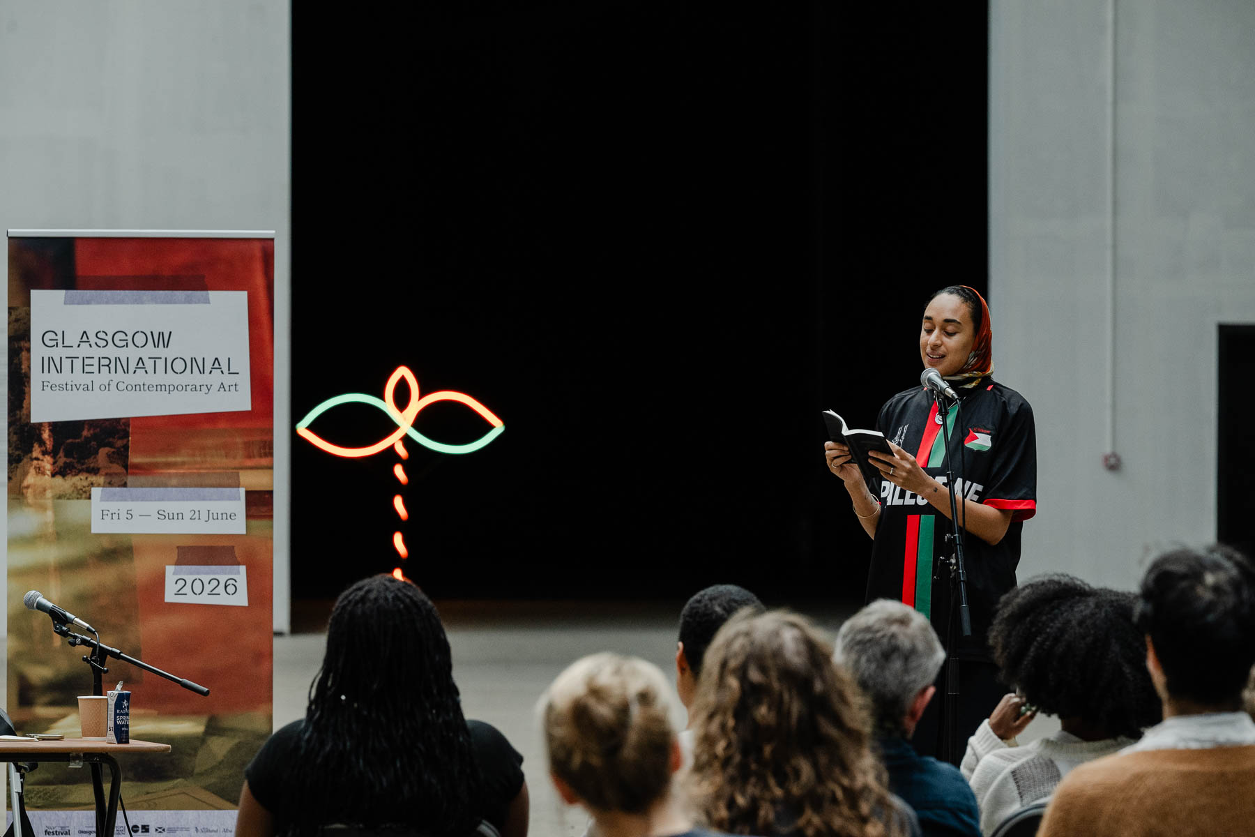 A woman wearing a headscarf and a "Palestine" football shirt stands reading from a book into a microphone in front of a seated audience, with a Glasgow International banner and a neon sculpture beside her.