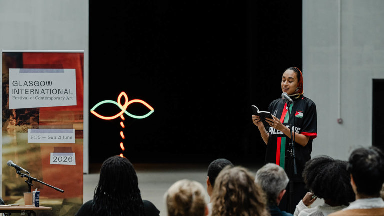 A woman wearing a headscarf and a "Palestine" football shirt stands reading from a book into a microphone in front of a seated audience, with a Glasgow International banner and a neon sculpture beside her.