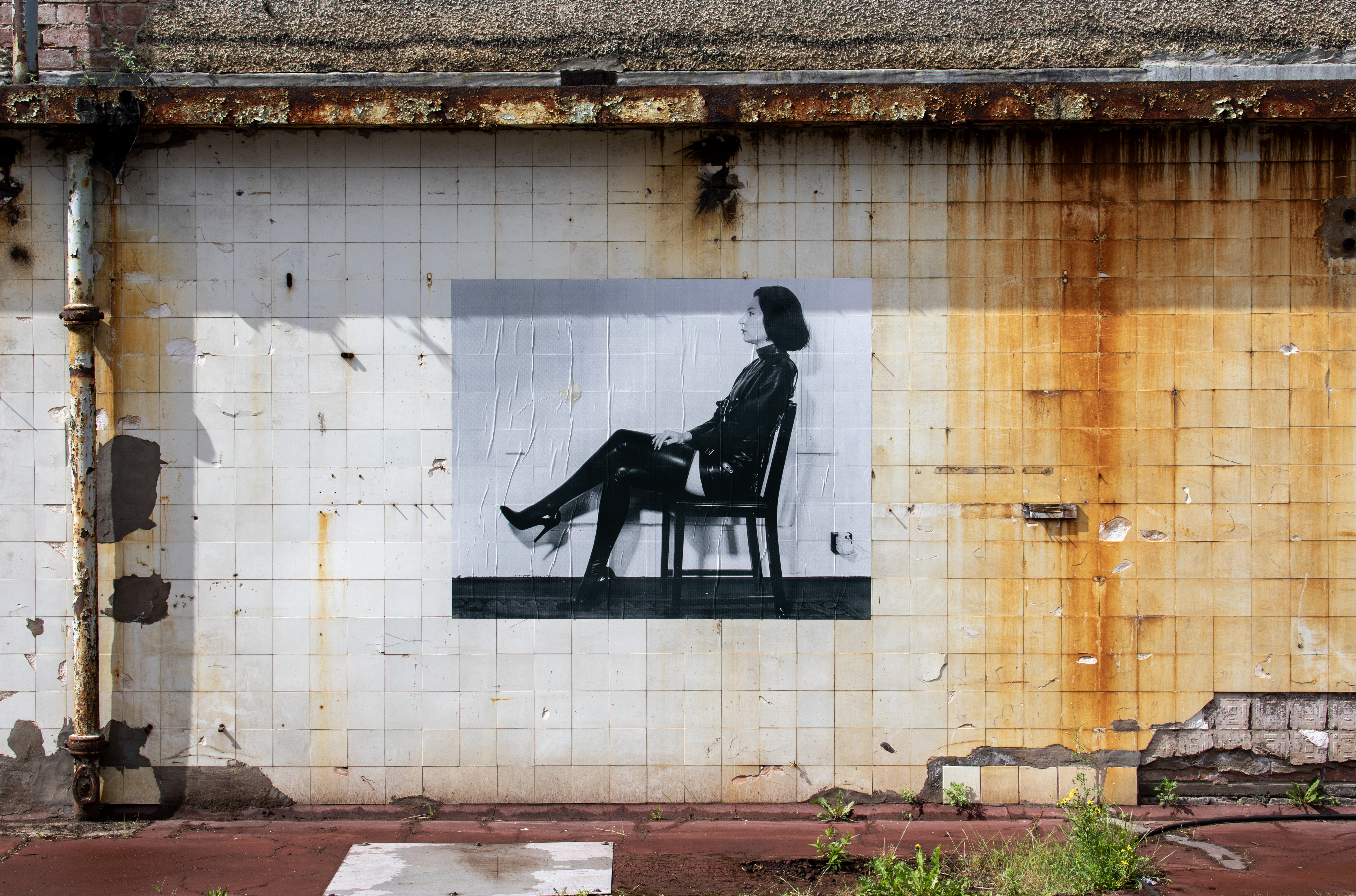 A weathered, rusted brick and tiled wall with a large black and white photograph pasted onto it, showing a woman in a black leather outfit seated sideways on a chair.