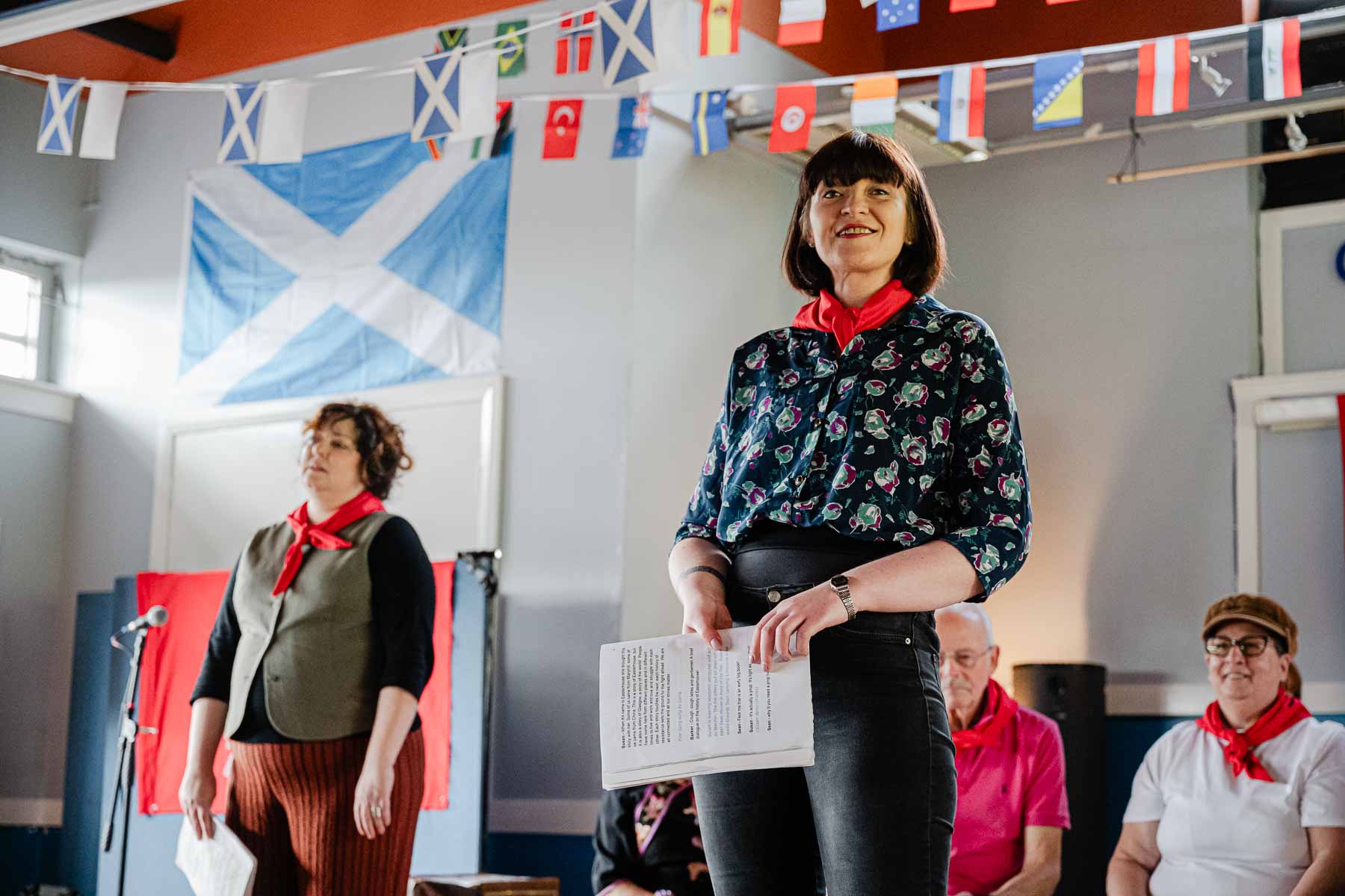 A performer in a floral blouse and red neckerchief smiles while holding a script, standing before a Scottish saltire flag, with another cast member behind her, part of Keira McLean's performance.