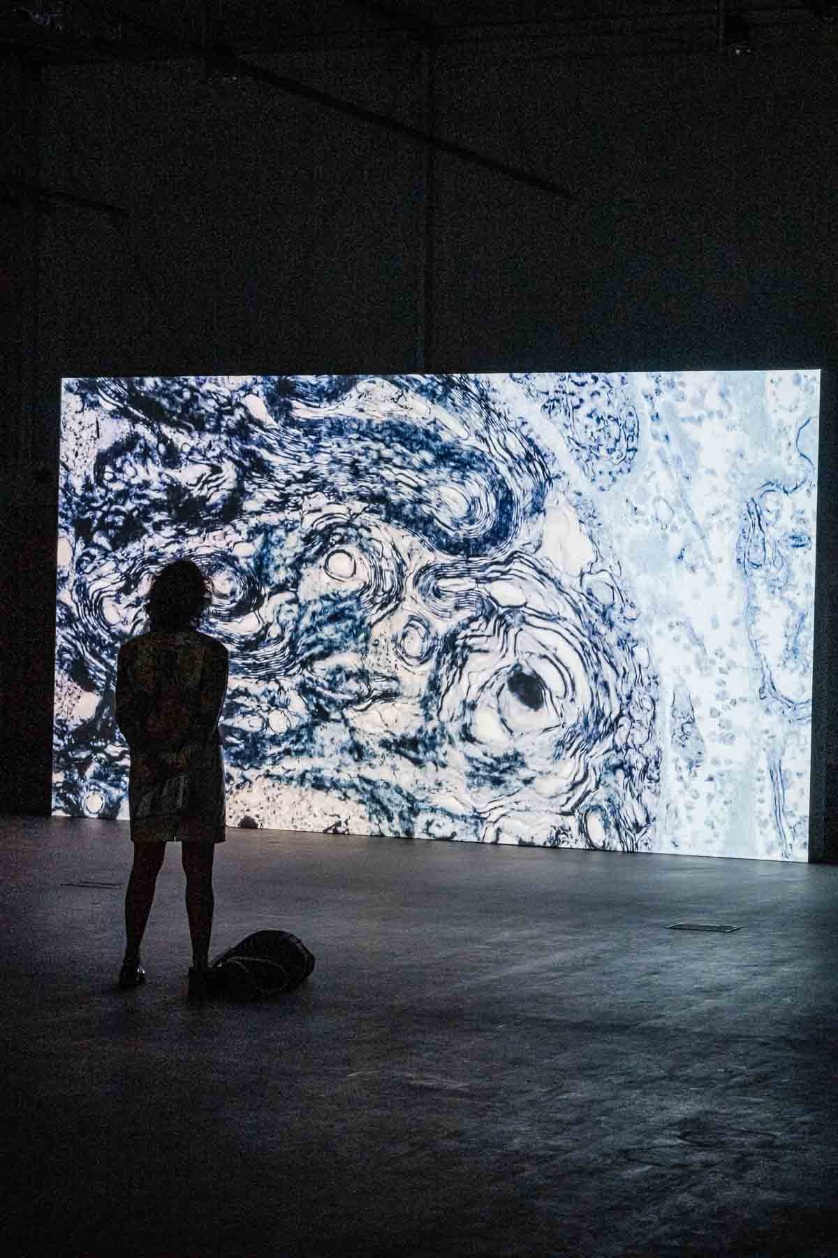 Installation view of a large video screen showing swirling abstract blue and white marbled patterns, with a person silhouetted in front of the projection.