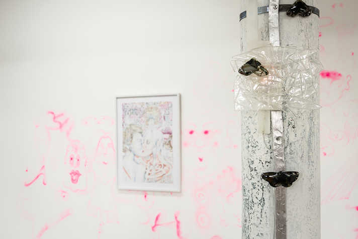 A framed artwork is displayed on a wall, it features two people kissing. In the foreground is a pipe which has black plastic butterflys attached to it.