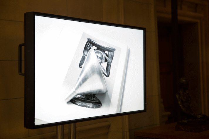 In a dimly lit corner of a room, there is a monitor placed on a stand, made of two metal poles. The monitor has a handle peeking behind it. On the screen is an image of a book with a monochromatic image of an ancient sculpture of an animal, holding a plate on its head. Placed on this book is a metallic funnel. The funnel is also monochromatic.