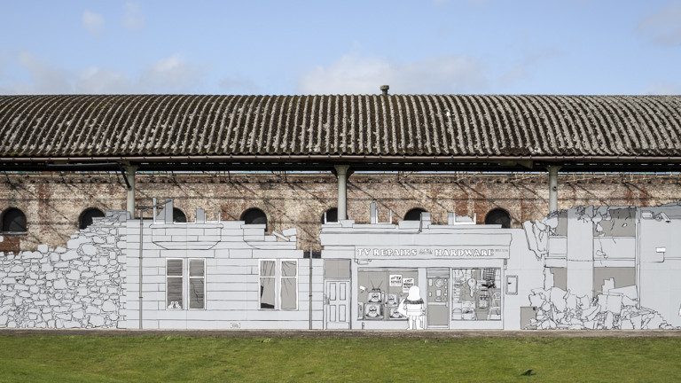 There is a blue sky with a few clouds. There is an old building and in front of it there is a black and white mural that resembles a street that depicts from left to right: a house, a TV repair and hardware store with someone looking at the window display and a building in ruins. The artwork has some green grass in front of it.