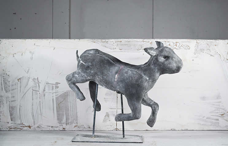 A cast aluminium lamb is posed leaping. The lamb has shiny markings across its body and a rough salty surface. There is a small pink gash in the side of the lamb. The lamb sits in front of a white painted wood background