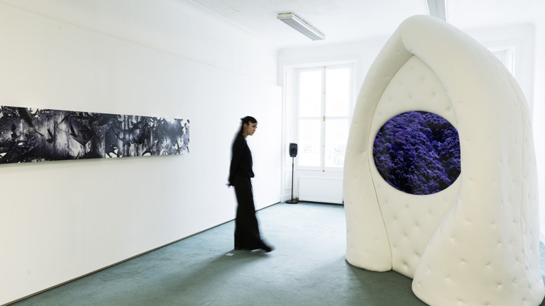 Installation view with a figure in dark, flowing clothing walking past a long black-and-white panel painting on the left wall. To the right, a large white padded, biomorphic arch-like sculpture has a rounded screen embedded in it, displaying a dense purple, tree-like or foliage image.
