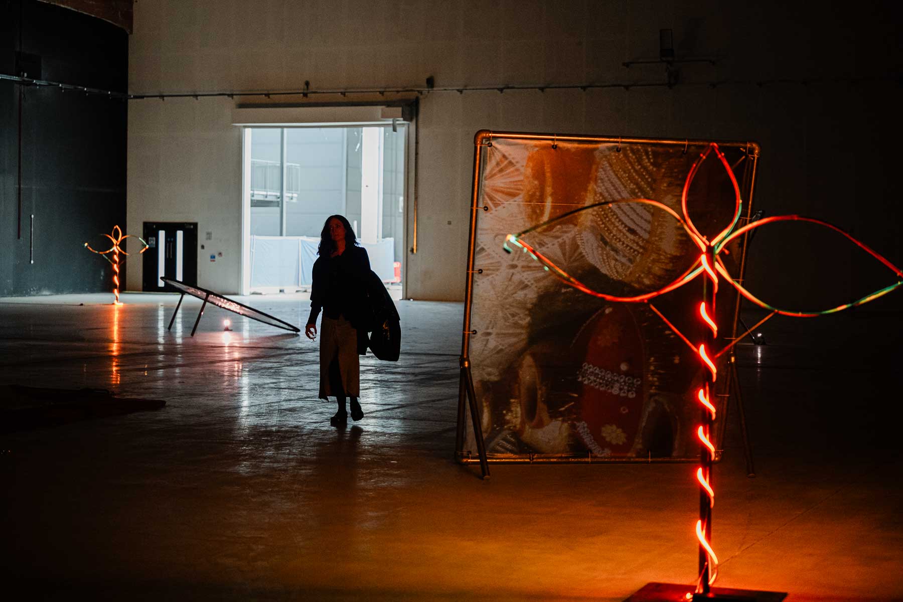 Installation view of a warehouse-style gallery with a large freestanding printed banner and neon plant-like sculptures glowing in orange and green, a figure walking through the dimly lit space towards a bright doorway in the background.