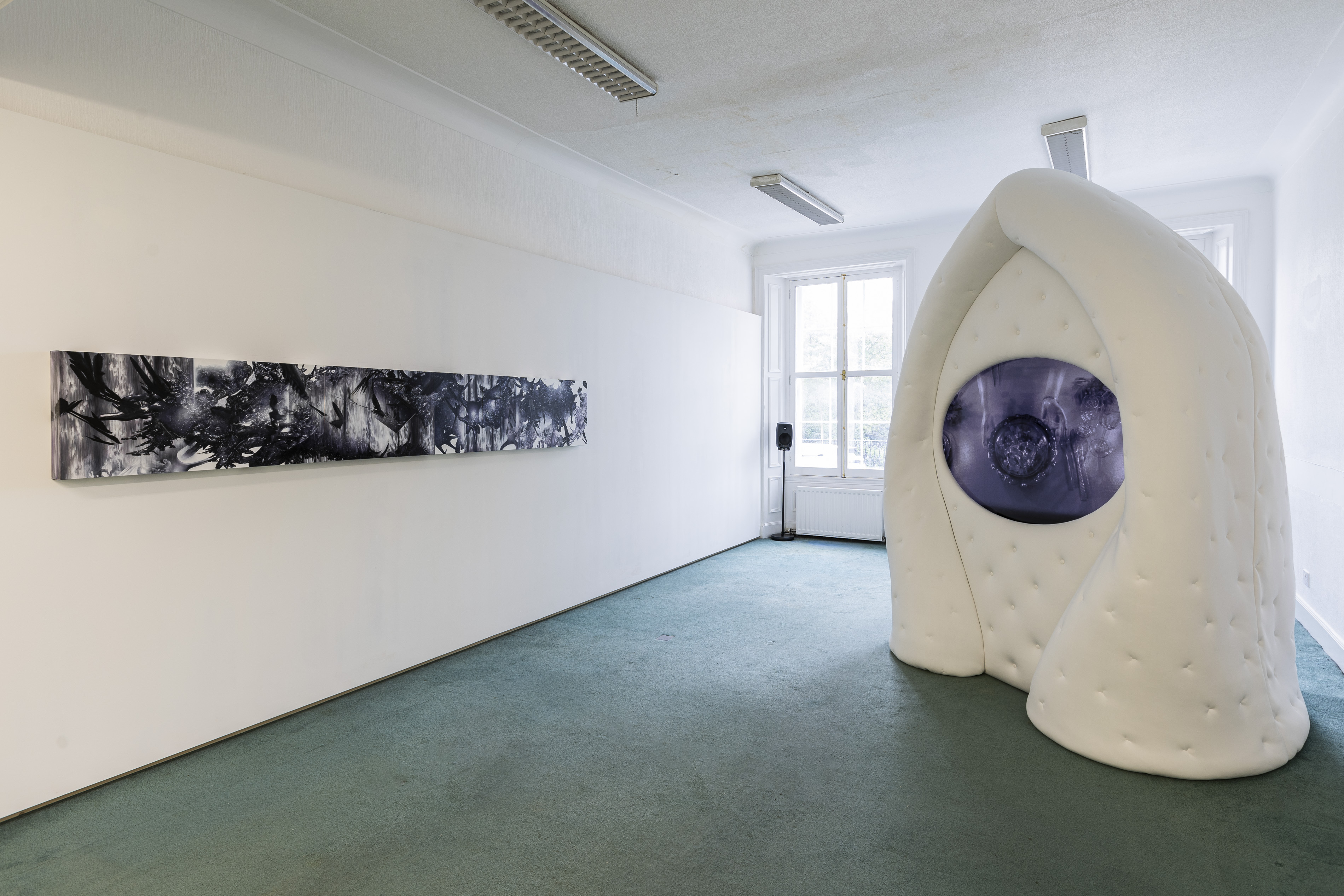 Installation view of a white gallery room with green carpet. On the left wall, a long horizontal painted panel shows abstract black and white forms suggesting birds and foliage. On the right, a large white padded, biomorphic arch-like structure stands in the room with a rounded video screen embedded in its surface, showing a swirling purple-toned image.