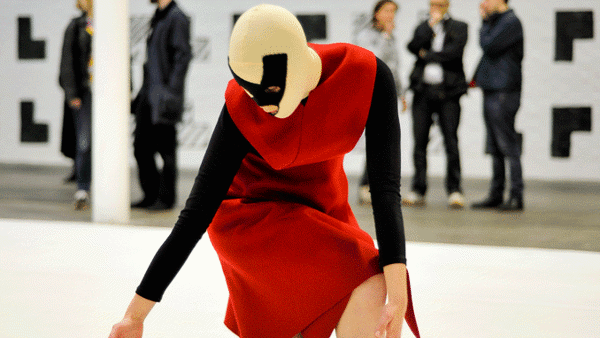 masked figure in red, black and white in dance pose within gallery space, individuals in the background blurred