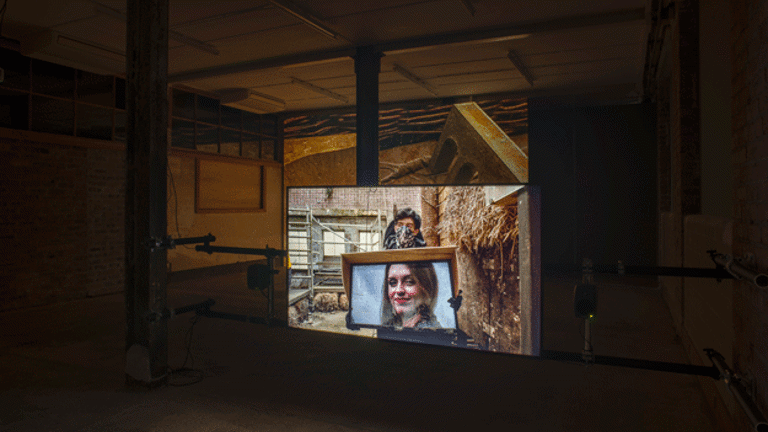 In a dark room a film is projected onto a screen. The projected image shows a light skinned person with dark hair and a facemask holding a tv screen with another light skinned, dark haired person with red lipstick smiling out from it