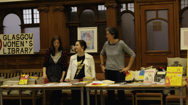 Three women are standing behind a line of tables. The table has multiple books and publications placed on it. One of the books reads the title "21 Revolutions". Behind the women, there is a poster saying "Glasgow Women's Library", a picture of a sculpture and a framed painting.