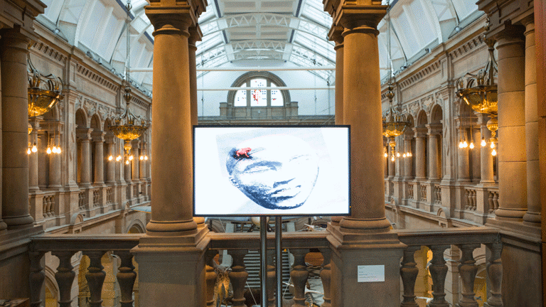 Giant arches and pillars are lit up by lights and hanging chandeliers. A long hall with tall arches running along it can be seen past the balcony. In front of the balcony, at the centre, there is a television screen standing on a stand made up of two metal poles. The screen has the image of photograph of a monochromatic stone mould of a face, on top of which sits a tiny bright coloured frog.