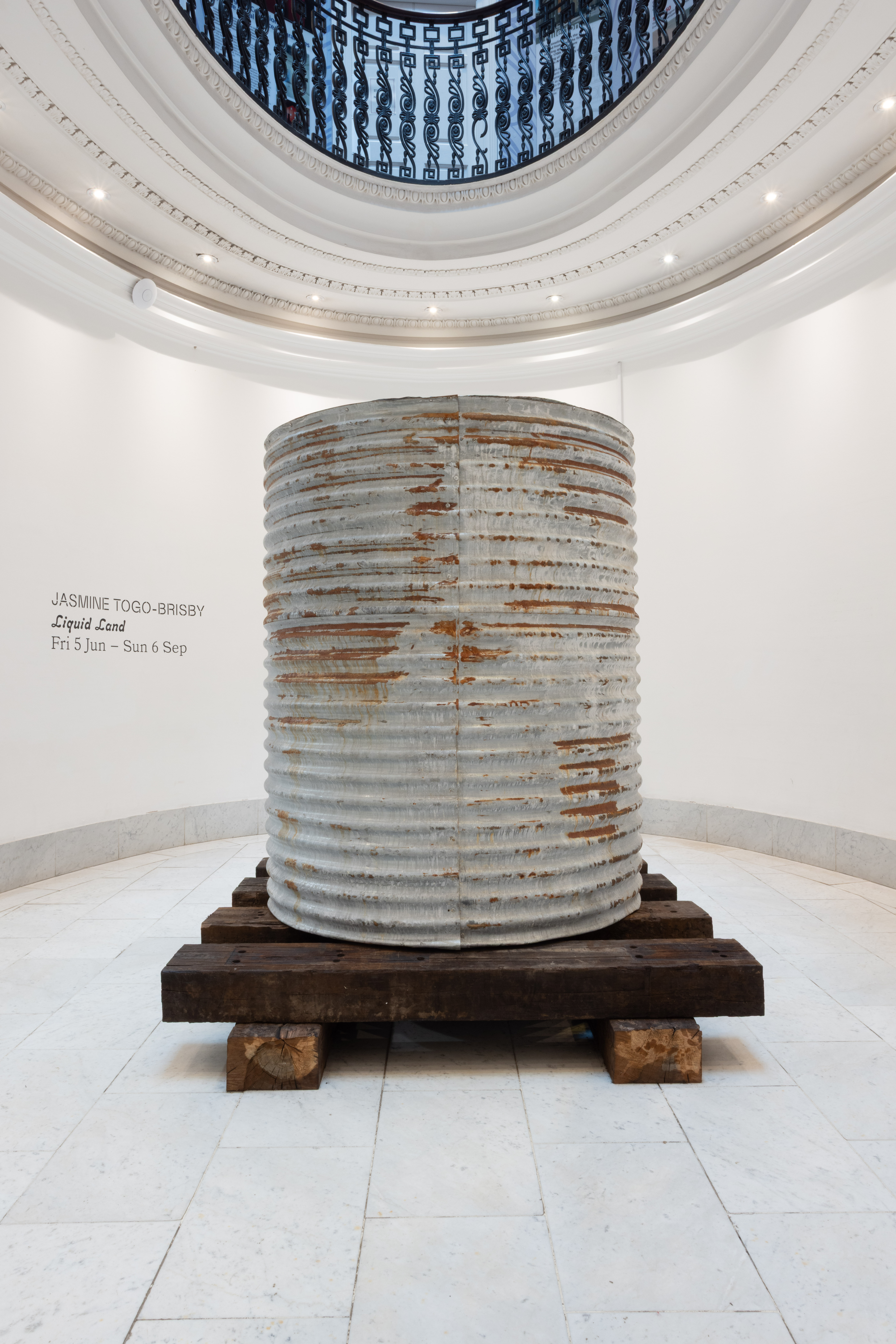 Installation view of a large corrugated metal cylindrical sculpture resting on timber beams beneath a domed skylight, with wall text reading "Jasmine Togo-Brisby, Liquid Land, Fri 5 Jun – Sun 6 Sep."