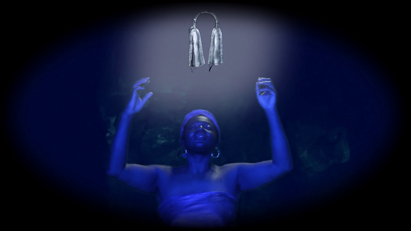 A woman, painted all in blue, raises her hands with her eyes closed underneath a bright light.