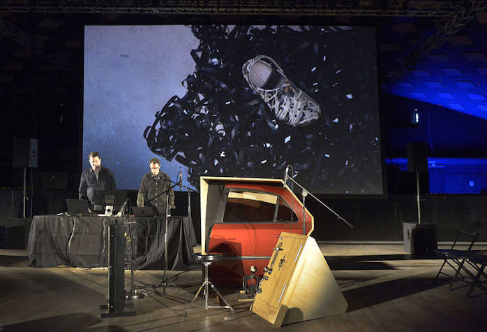 there are two people stood in front of a desk with laptops. behind them is a screen with the image of a croc shoe on top of tape reel. in front of them is a re car door framed by plywood and surrounded by microphones
