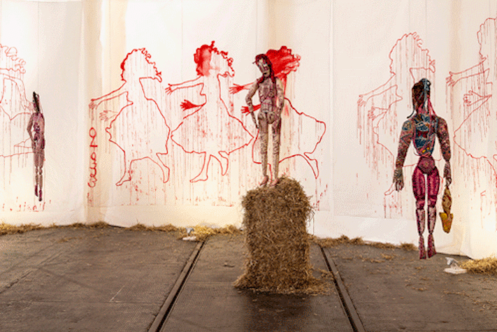 Drawings and artwork on fabric hanging from a wall. In front of the fabric is a sculpture of a figure on a straw bale.