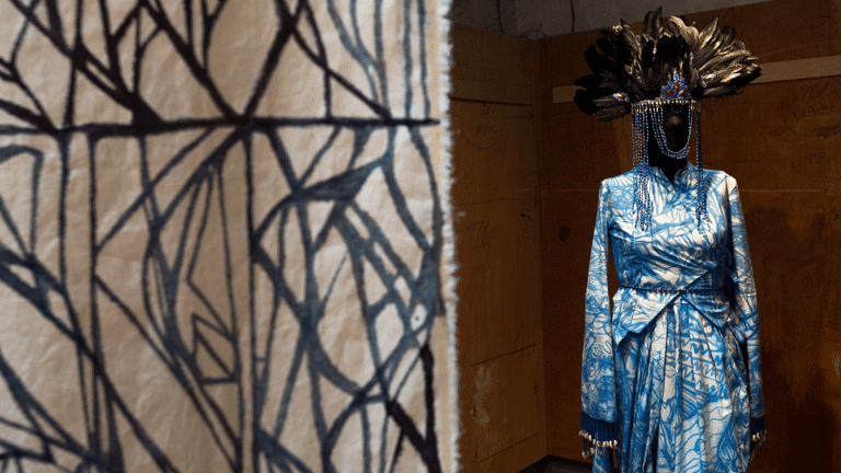 In the foreground a batic hanging on calico. In the background a manequin with an intricate dress made from fabric with an abstract indigo painted pattern. The manequin wears a headdress of large black feathers with strands of beads draped around the face.