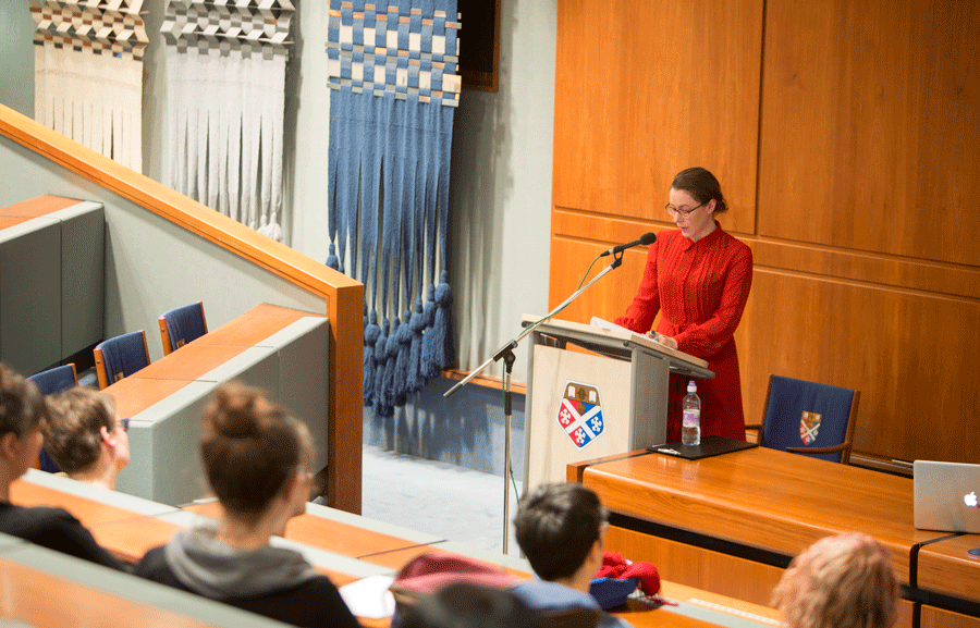 In a lecture theatre, a woman is giving a speech, through a microphone, standing behind a podium. There are many people sitting in front of her on benches, in the lecture theatre.
