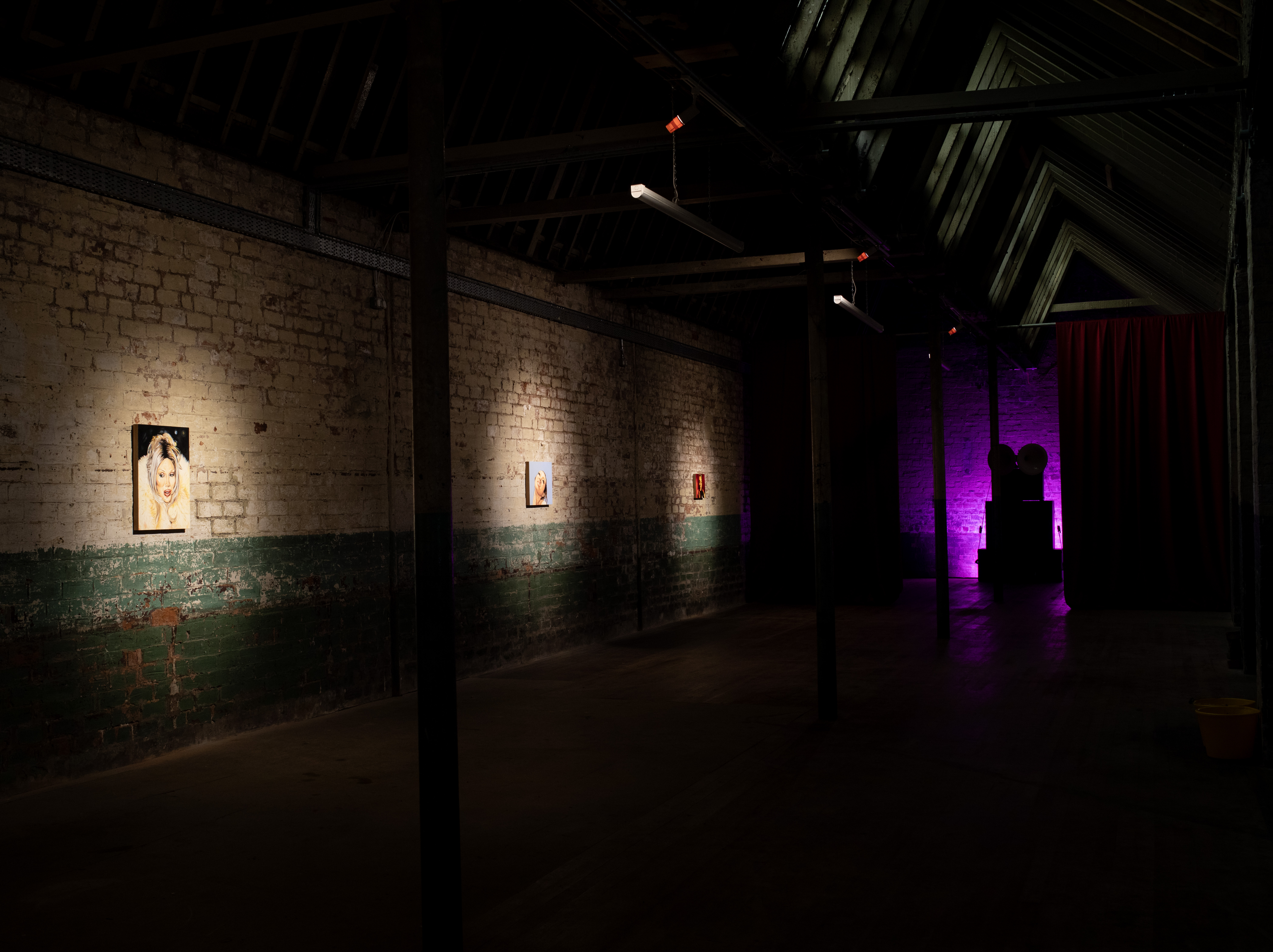 Dim gallery corridor with exposed brick walls, green-painted lower half. Three small spotlit portrait paintings hang in sequence along the left wall; a purple-lit space with dark silhouetted objects and a red curtain is visible in the background.