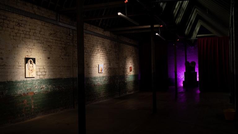 Dim gallery corridor with exposed brick walls, green-painted lower half. Three small spotlit portrait paintings hang in sequence along the left wall; a purple-lit space with dark silhouetted objects and a red curtain is visible in the background.
