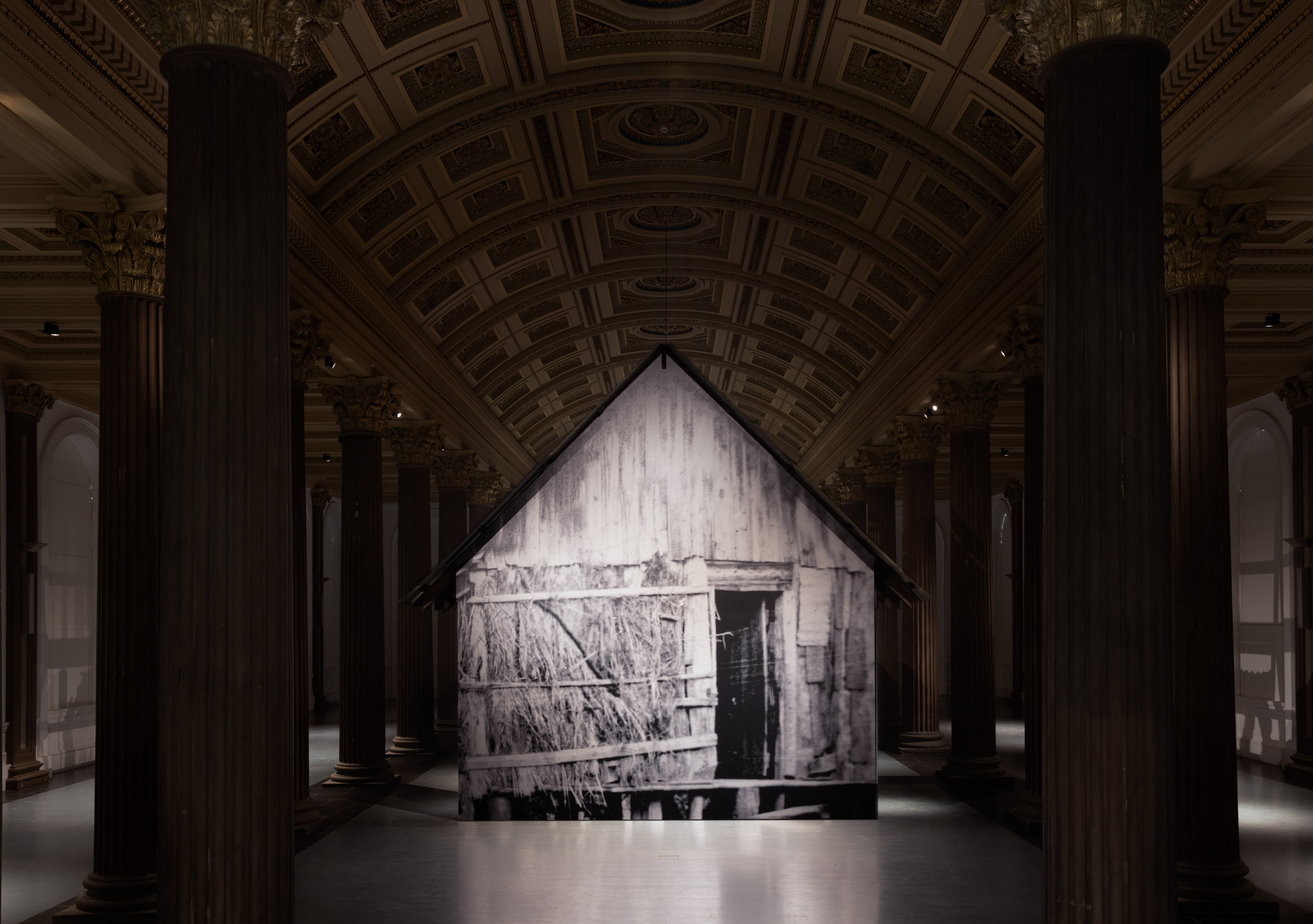 Installation view of a large freestanding structure printed with a black and white archival image of a timber and thatch hut, suspended within a grand columned hall with an ornate coffered ceiling.