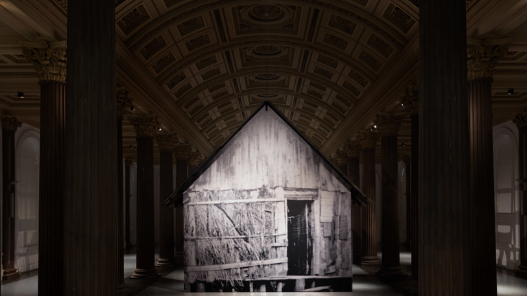 Installation view of a large freestanding structure printed with a black and white archival image of a timber and thatch hut, suspended within a grand columned hall with an ornate coffered ceiling.