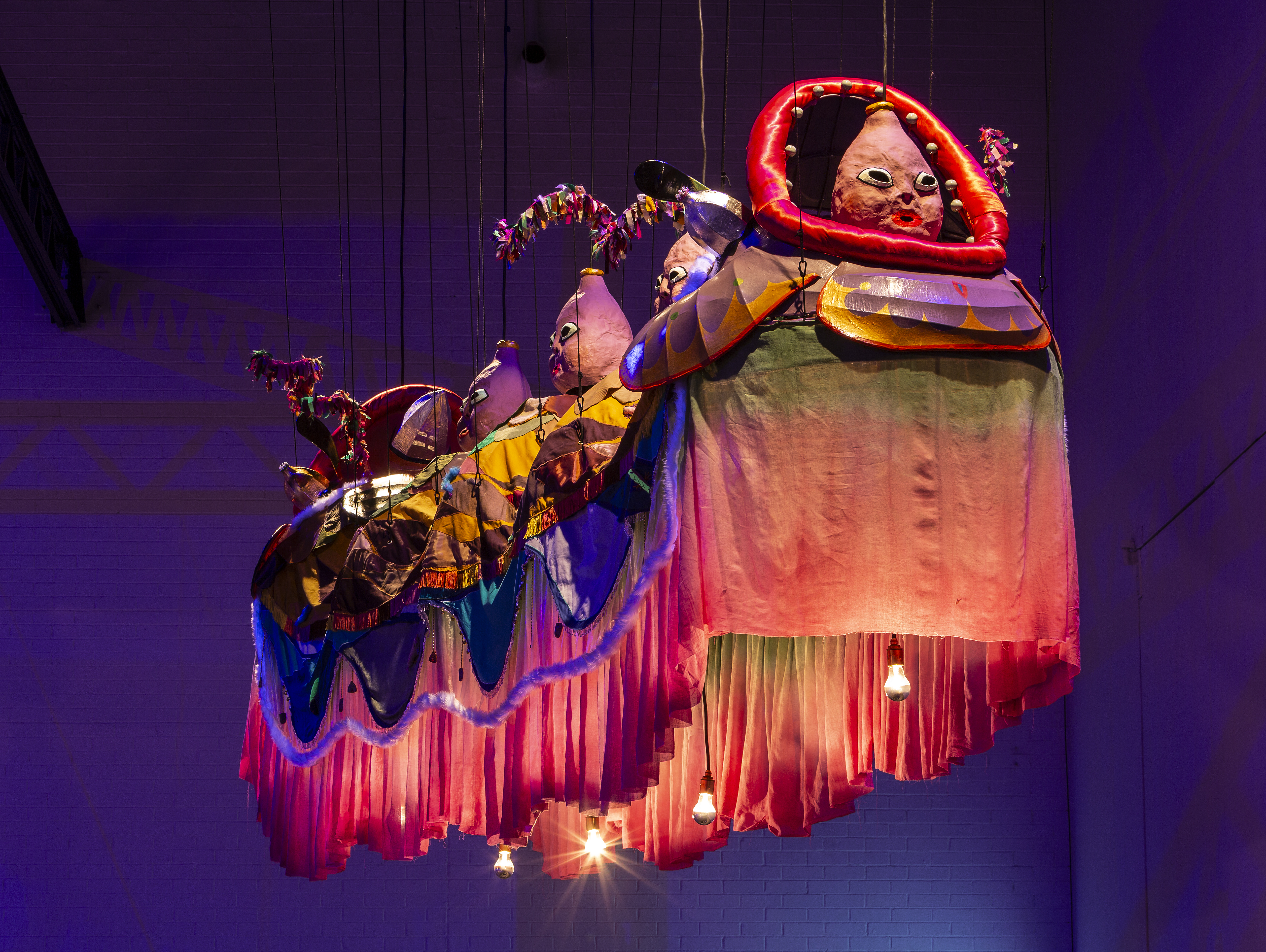 A suspended sculptural piece. There is a range of materials including fabric, paper mache, and lighting. The papermache displays vase like heards that run throughout the body of the work. Under these heads is fabric draping of pink and blue. 