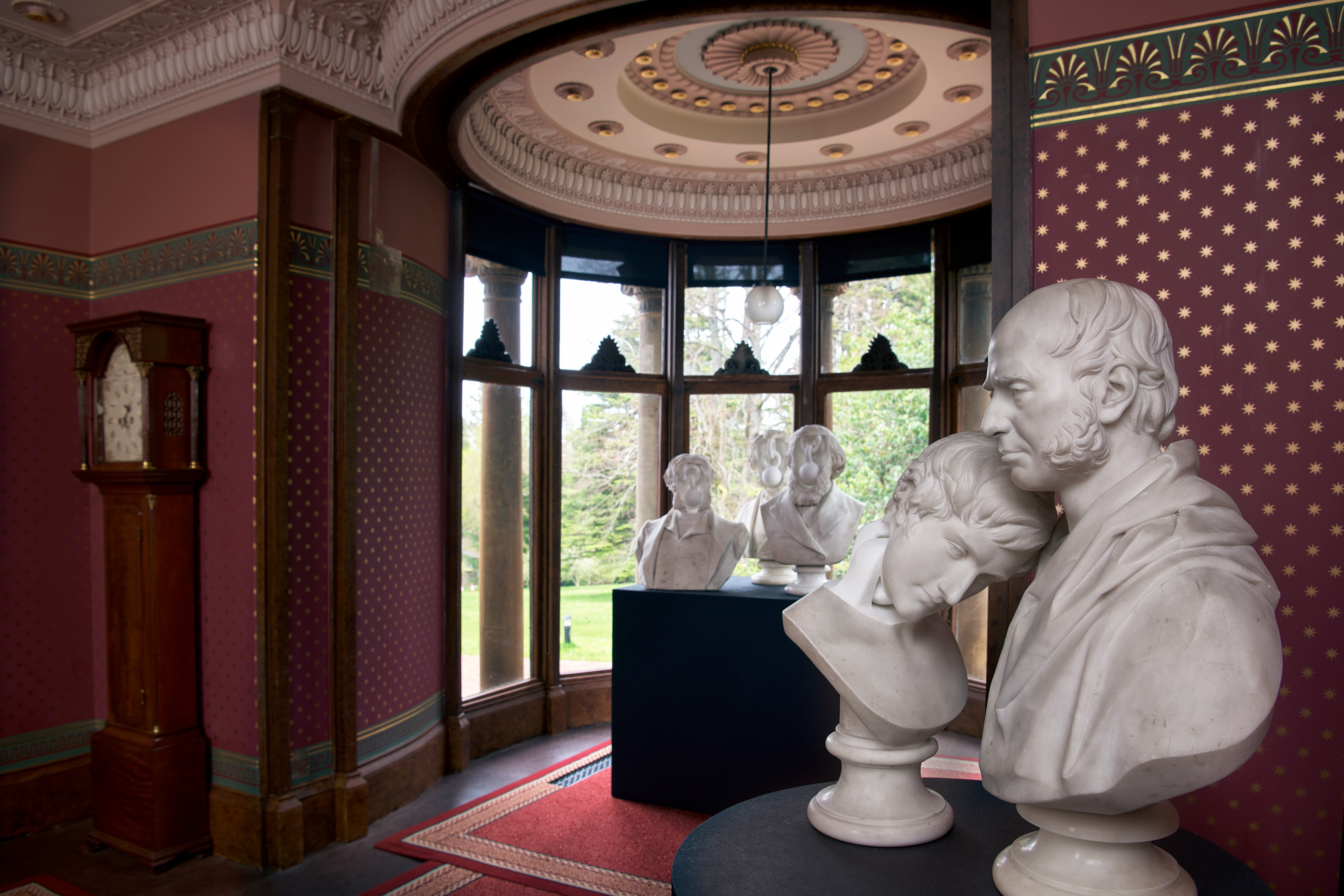 A close-up of a white marble double bust, two heads leaning together, set against Holmwood House's painted red frieze wall, part of Jon Owen's installation.