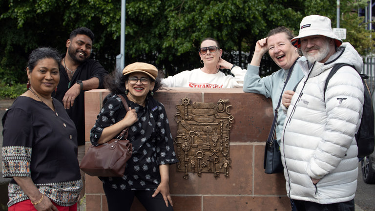 A group of six people laugh together outdoors around a stone plinth bearing a bronze relief of keys, tools, and small objects, part of Mandy McIntosh's sculpture at Springburn.