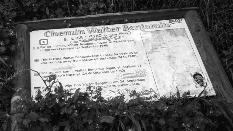 A black-and-white image, a plaque covered by foilage has the text "Chemin Walter Benjamin"