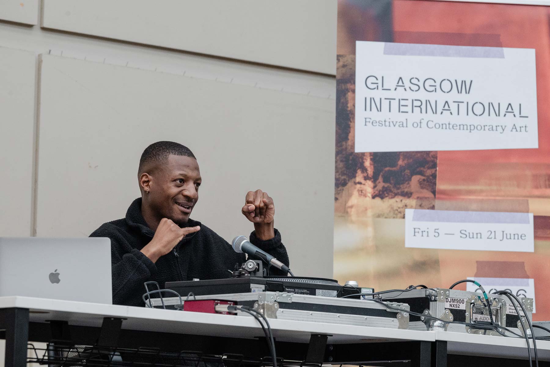 Ashley Holmes gestures while speaking at a table with a laptop and DJ mixer in front of him, a microphone positioned nearby. A Glasgow International banner is visible behind him.